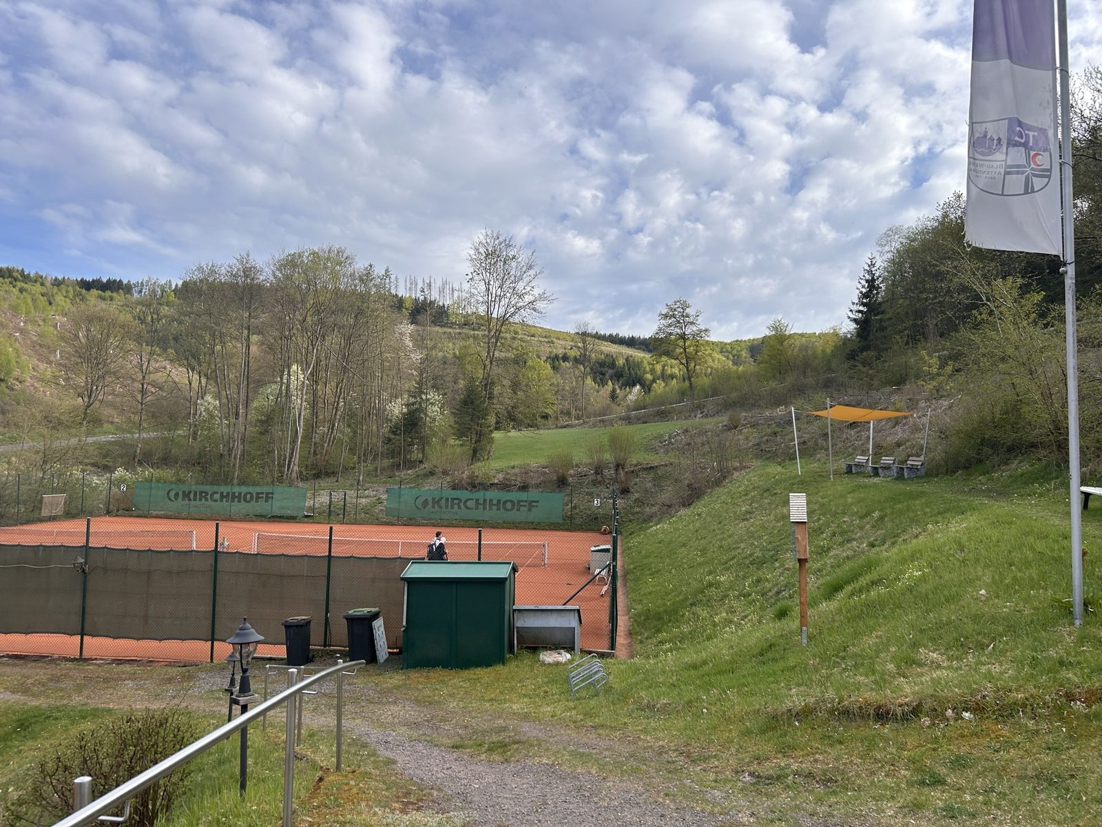 Blick über die Tennisanlage mit Vereinsfahne und Hang im Hintergrund